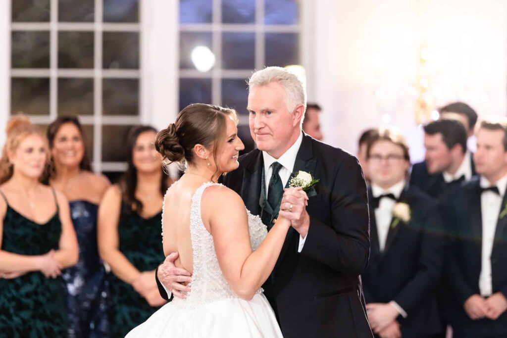 Bride sharing an emotional father-daughter dance with her dad during the wedding reception, surrounded by their bridal party in heartfelt admiration.