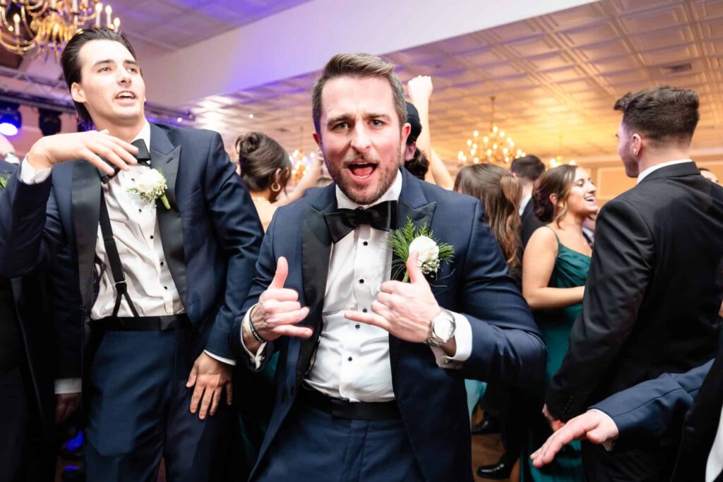 Groomsman in a navy tuxedo dancing and making playful gestures during a lively wedding reception, surrounded by guests enjoying the celebration.