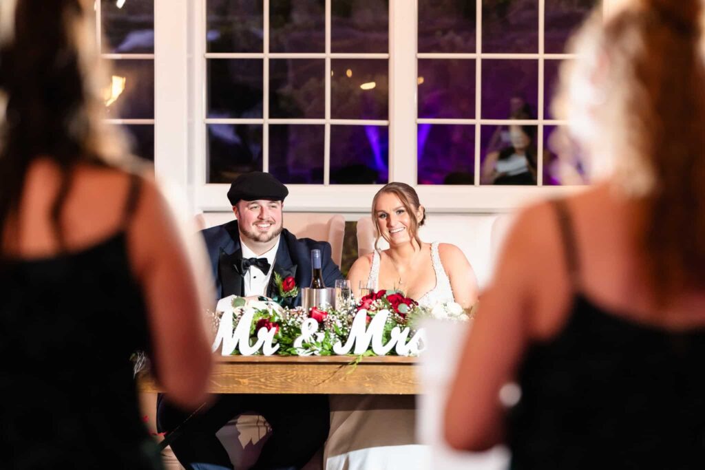 Bride and groom seated at their sweetheart table adorned with “Mr. & Mrs.” sign and florals, smiling during a heartfelt wedding toast.