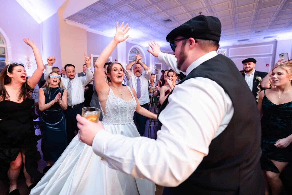 Bride and groom celebrating on the dance floor with arms raised, surrounded by cheering wedding guests during a joyful winter reception.