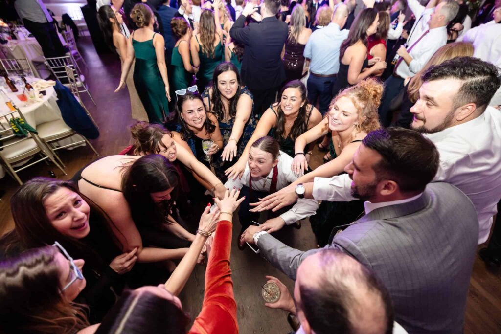 Wedding guests forming a joyful huddle on the dance floor, reaching hands in together during a spirited moment of celebration at the reception.