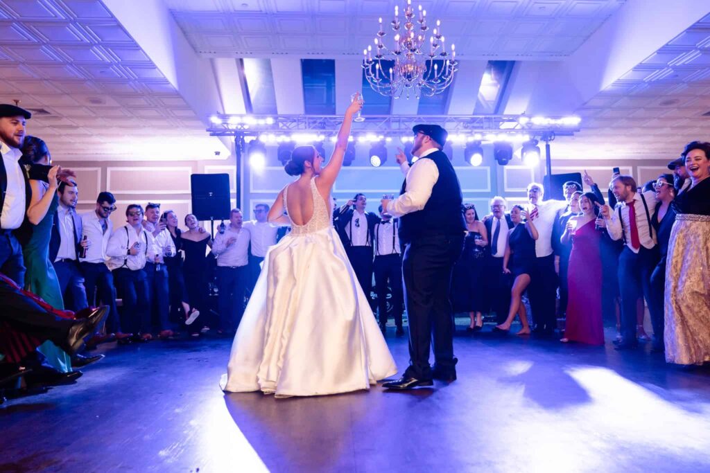 Bride and groom raising glasses on the dance floor at East Wind, surrounded by cheering guests under vibrant lighting and a sparkling chandelier during their wedding reception.