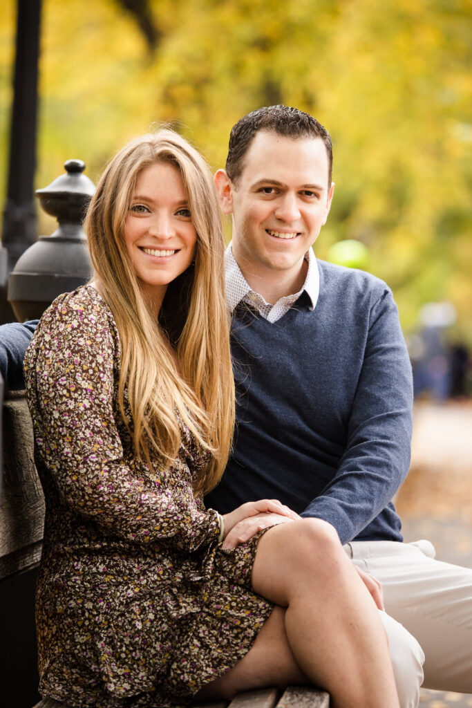 Engaged couple sitting on a bench in during their engagement session, smiling with golden autumn leaves in the background.