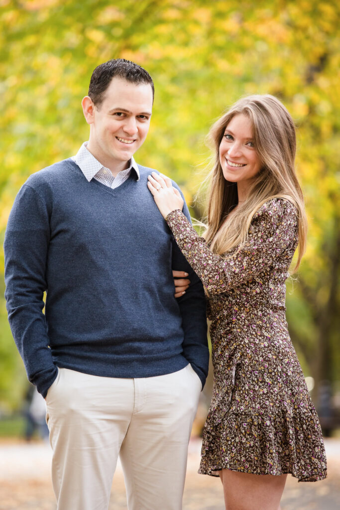 Couple posing together under vibrant foliage, with the bride-to-be’s hand resting on her fiancé’s shoulder during their autumn engagement session.
