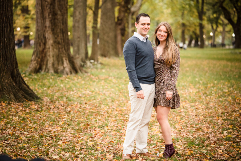 Engaged couple standing on a leaf-covered lawn in Central Park during peak fall color, surrounded by tall trees and golden light in their NYC engagement session.