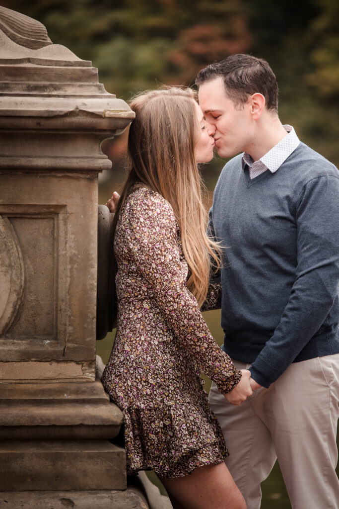 Couple sharing a quiet kiss beside the stone details of Bethesda Fountain during their engagement session, holding hands with soft autumn tones in the background.