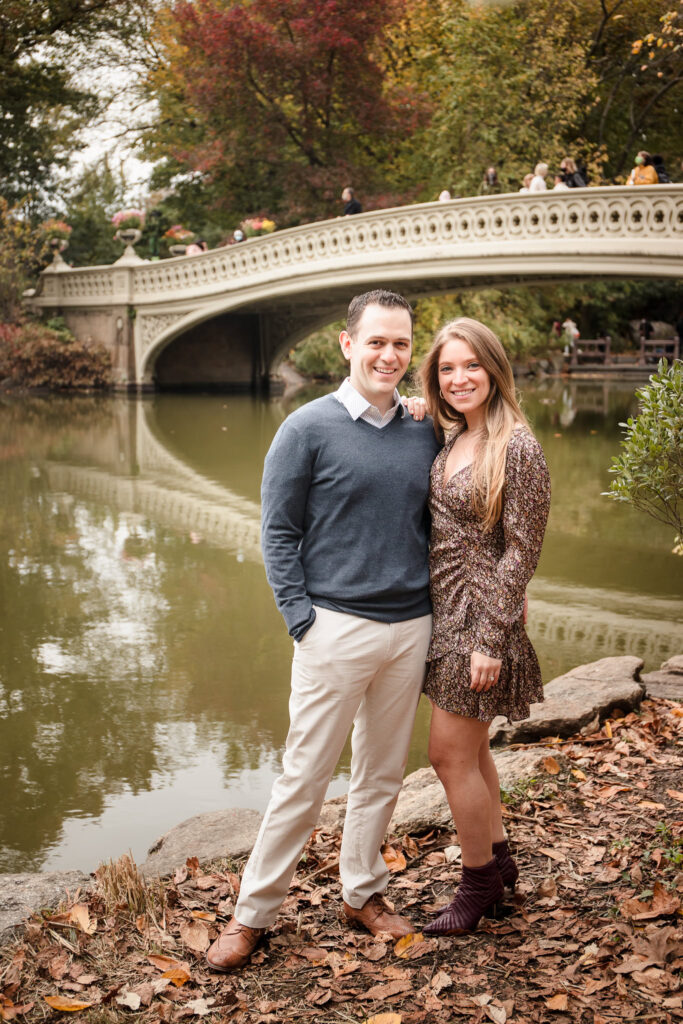 Engaged couple standing near Bow Bridge in Central Park, surrounded by fall leaves and colorful foliage during their autumn engagement photo session in NYC.