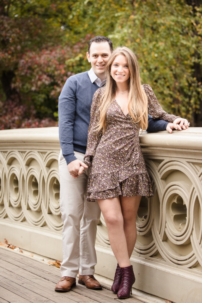 Couple holding hands and smiling while standing on Bow Bridge during their engagement session in Central Park, surrounded by soft foliage and classic architecture.
