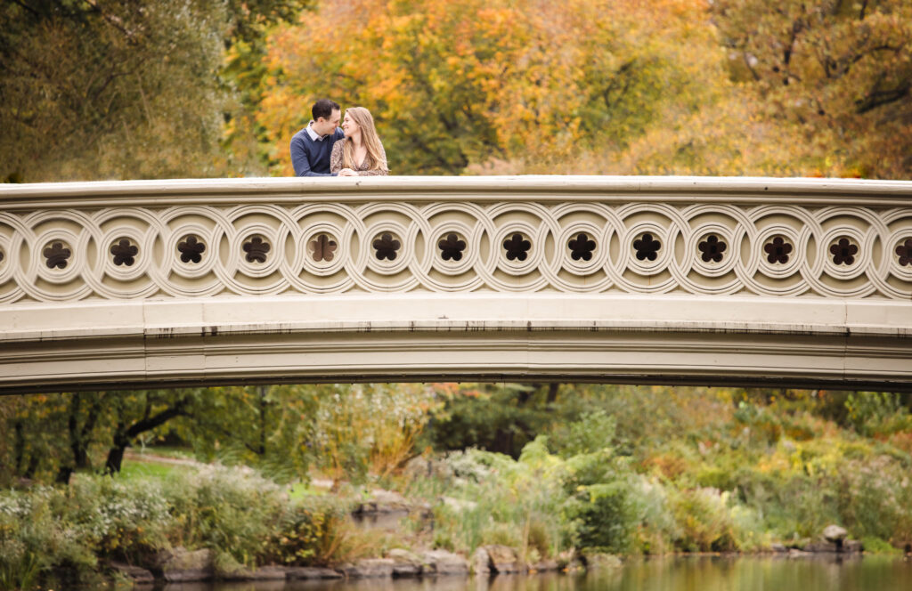 Wide view of engaged couple leaning on Bow Bridge in Central Park, surrounded by peak fall foliage and soft reflections during their NYC autumn engagement session.