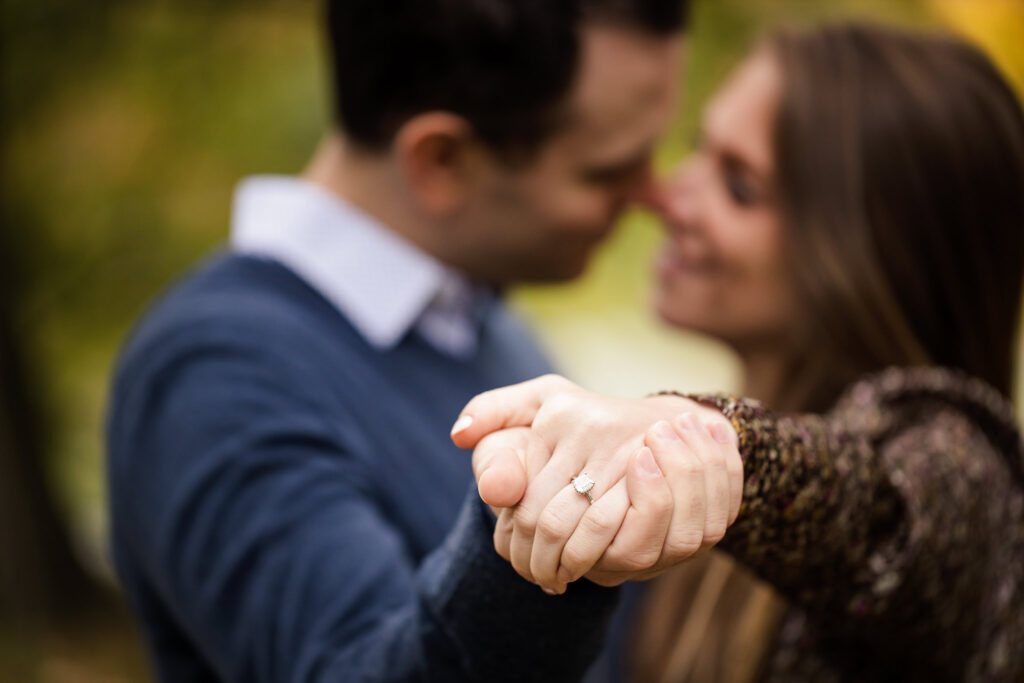 Close-up of bride-to-be’s hand showing her engagement ring as the couple embraces in soft focus during their engagement session.