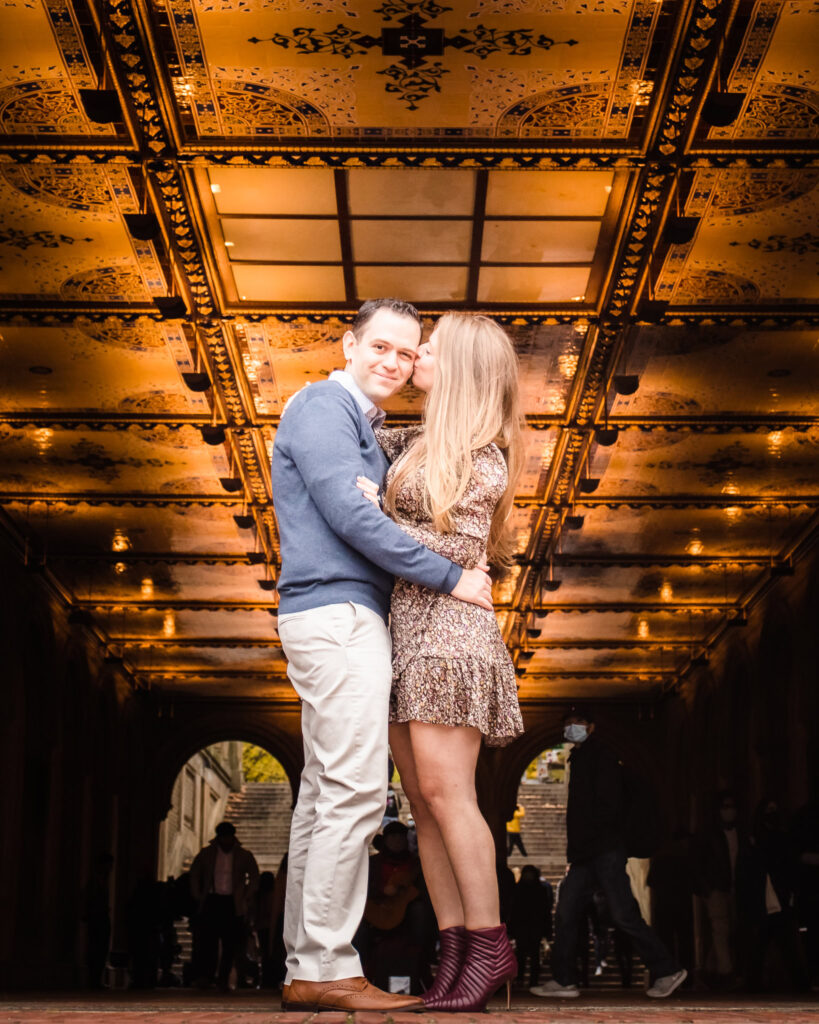 Bride-to-be kisses her fiancé’s cheek beneath the glowing Minton tile ceiling at Bethesda Terrace during their romantic fall engagement session in Central Park. All 15 alt texts are now