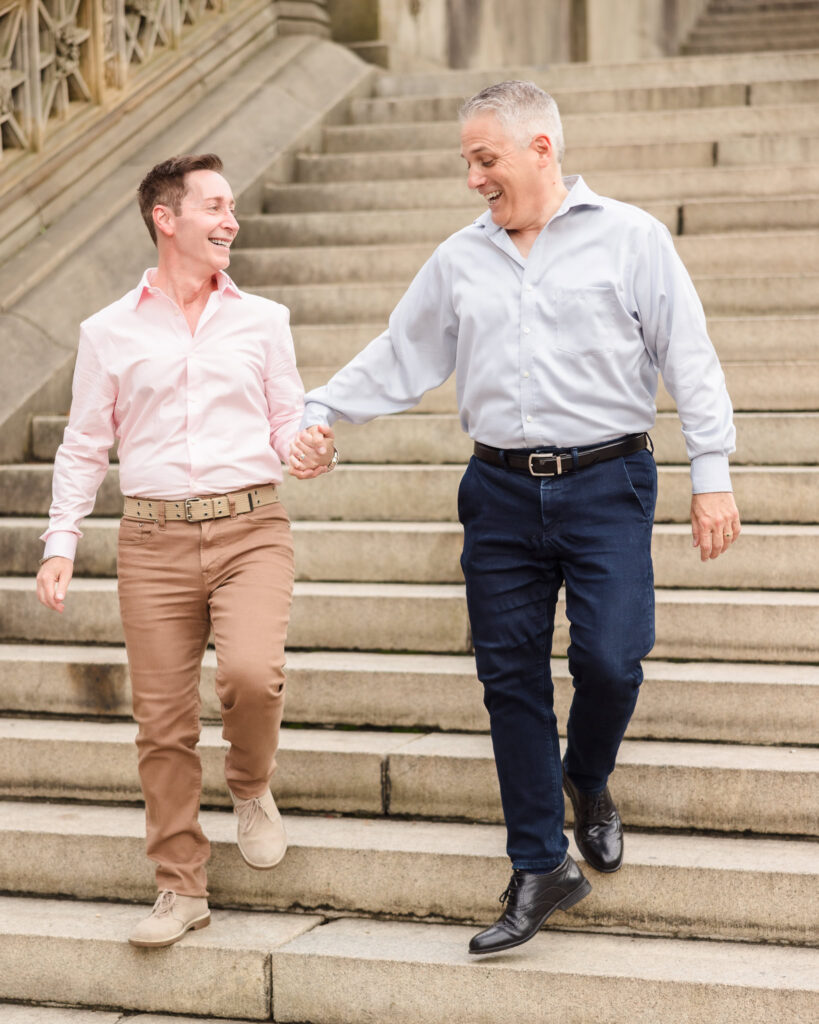Same sex couple walking hand-in-hand down the Bethesda Terrace steps in Central Park during their engagement session, smiling and sharing a joyful, relaxed moment together.