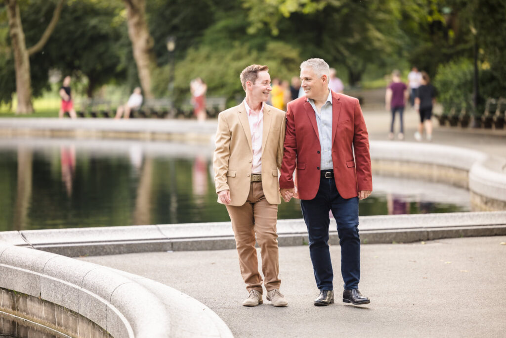 Engaged same sex couple walking hand-in-hand along the Conservatory Water in Central Park, dressed in tailored jackets and sharing a quiet, connected moment in the heart of NYC.