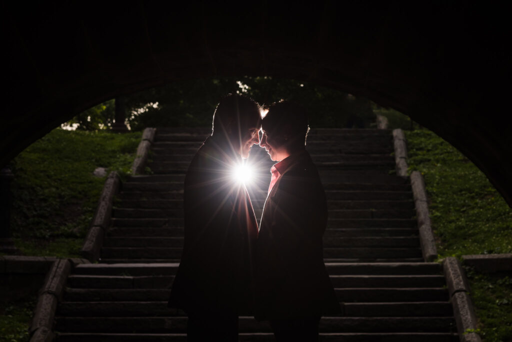 Silhouetted same sex couple touching foreheads under a Central Park bridge, with a soft burst of light between them creating a dramatic and intimate engagement portrait.
