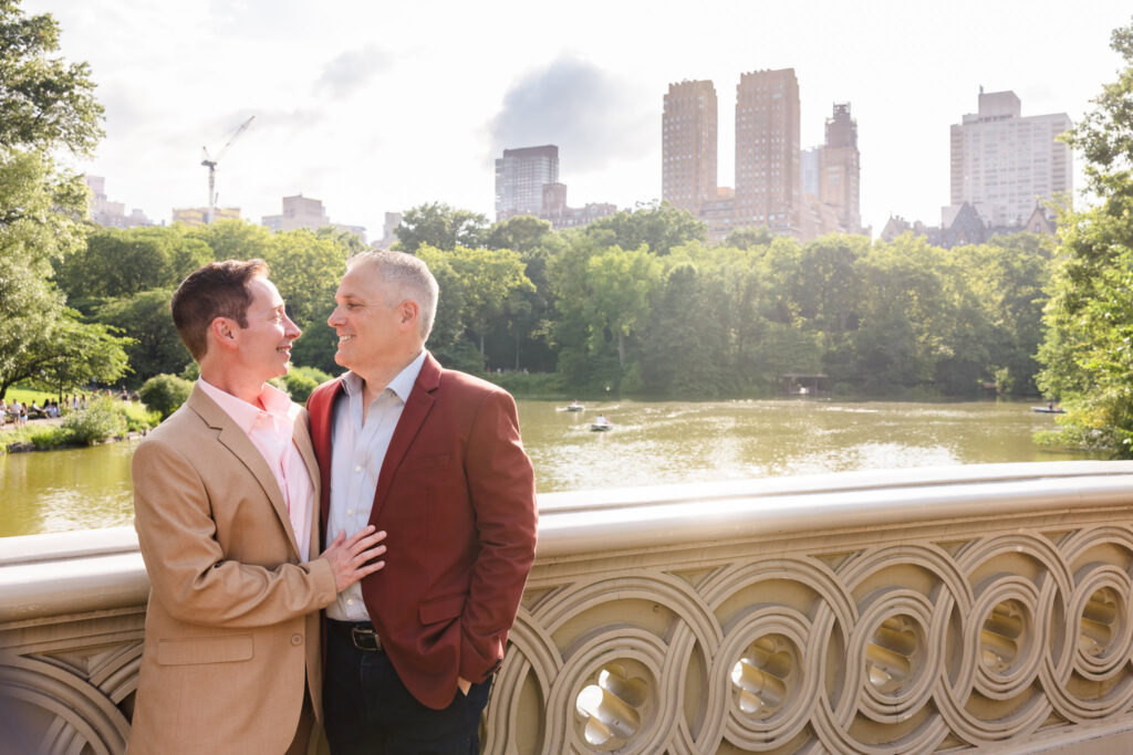 Same sex couple standing on Bow Bridge in Central Park during their engagement session, smiling at each other with the Manhattan skyline and The Lake in the background.