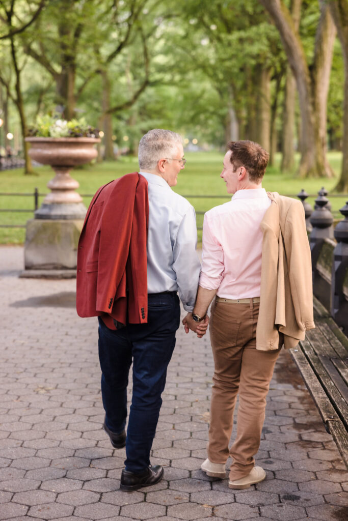 Engaged same sex couple walking hand-in-hand through The Mall in Central Park, jackets draped over their shoulders as they share a quiet, candid moment from behind.