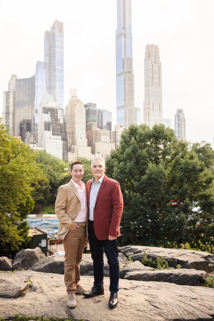 Engaged couple standing on a rocky outcrop in Central Park with the Manhattan skyline rising behind them, dressed in tailored jackets and smiling during their same sex engagement session.