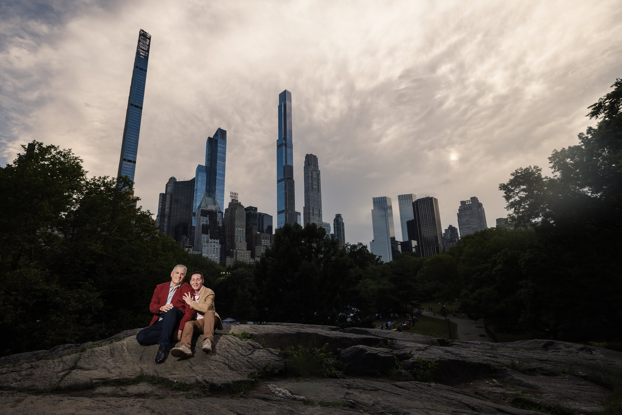 Same sex couple seated on a rocky ledge in Central Park, showing off their engagement rings with the dramatic Manhattan skyline and evening sky behind them.
