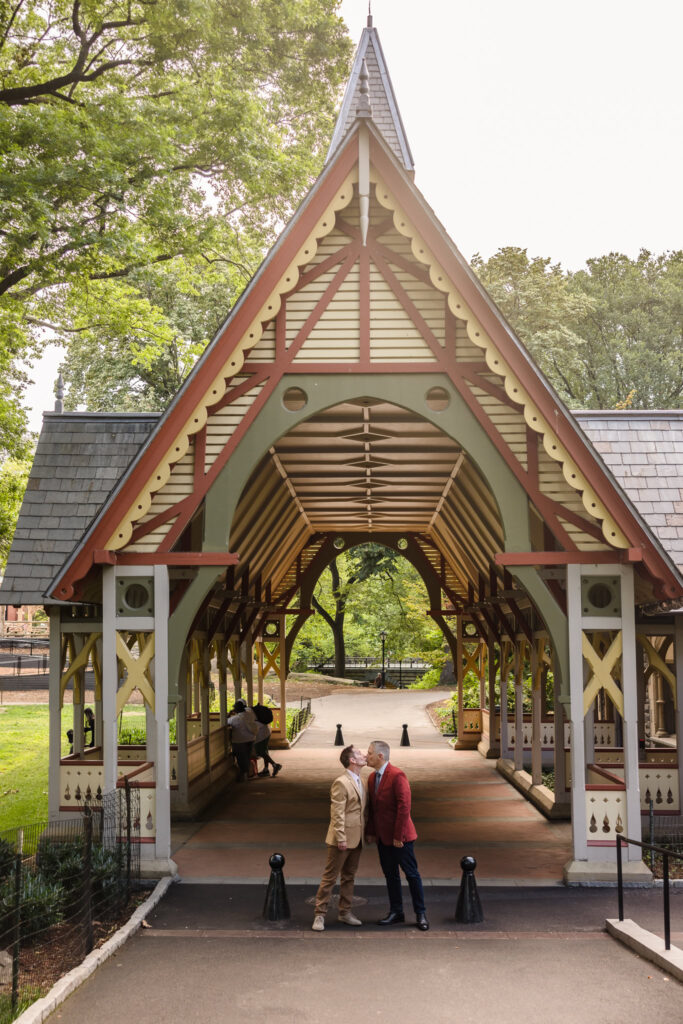 Same sex couple sharing a kiss beneath the colorful Victorian Gothic architecture of the Dairy Visitor Center in Central Park during their NYC engagement session.