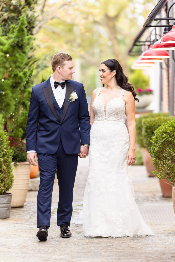 Bride and groom walk hand in hand outside Tellers Next Door venue in Islip, NY, surrounded by lush greenery and red pendant lights, a candid moment of connection on their elegant Long Island wedding day.