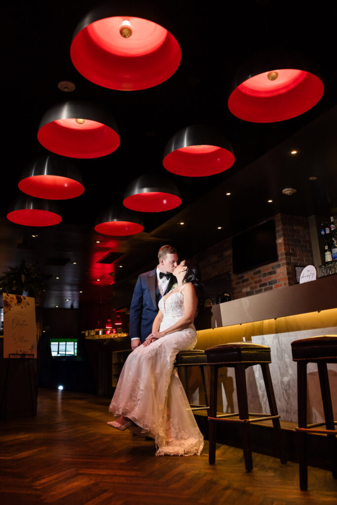 Bride and groom share a romantic kiss at the bar inside Tellers Next Door in Islip, NY, beneath dramatic red pendant lighting an intimate, stylish moment reflecting the venue’s modern elegance.