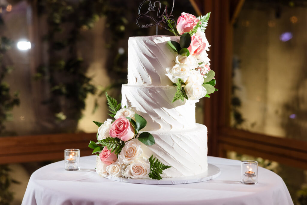 Three-tier white wedding cake adorned with blush and ivory roses, set on a round table with candlelight, a refined and romantic detail from an intimate Long Island celebration.