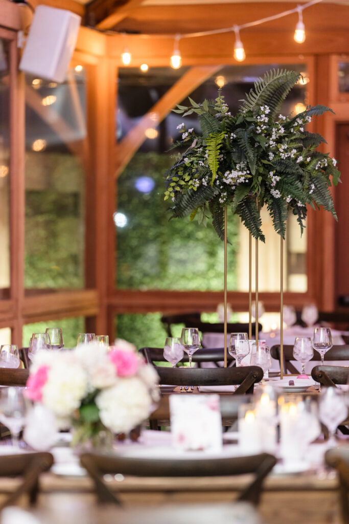 Close-up of a tall fern and floral centerpiece at a wedding reception, with soft candlelight, blush accents, and string lights creating a warm, inviting atmosphere in Islip, NY.