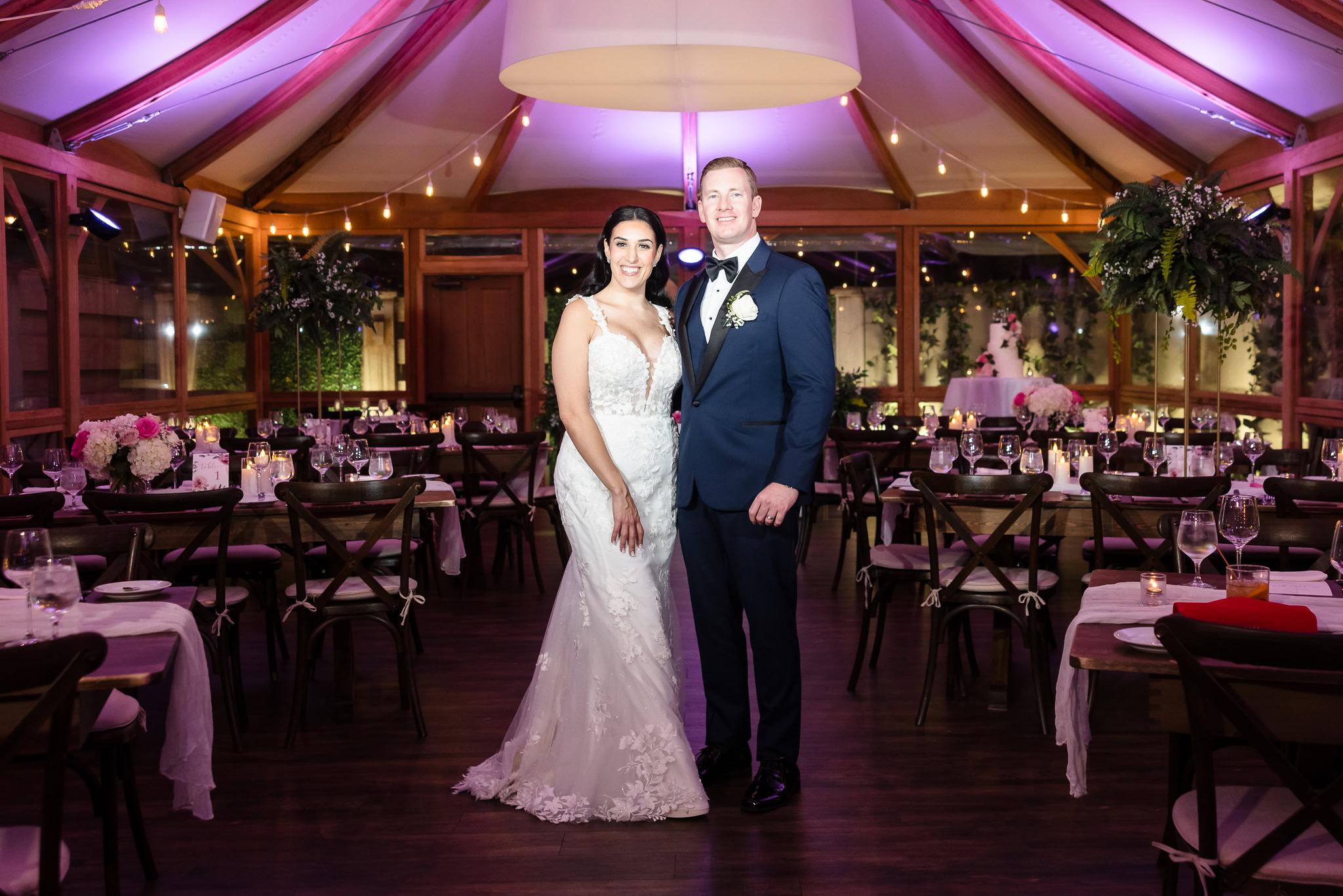 Bride and groom pose inside the Pavilion at Tellers Next Door, surrounded by candlelit tables, floral centerpieces, and romantic up lighting a beautifully styled intimate wedding reception in Islip, NY.