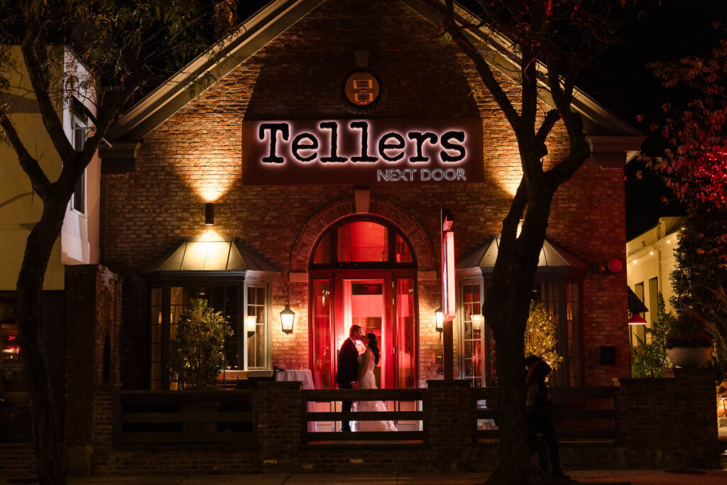 Exterior night view of Tellers Next Door in Islip, NY, with the bride and groom silhouetted in the glowing entrance, a romantic and cinematic moment at this intimate Long Island wedding venue.