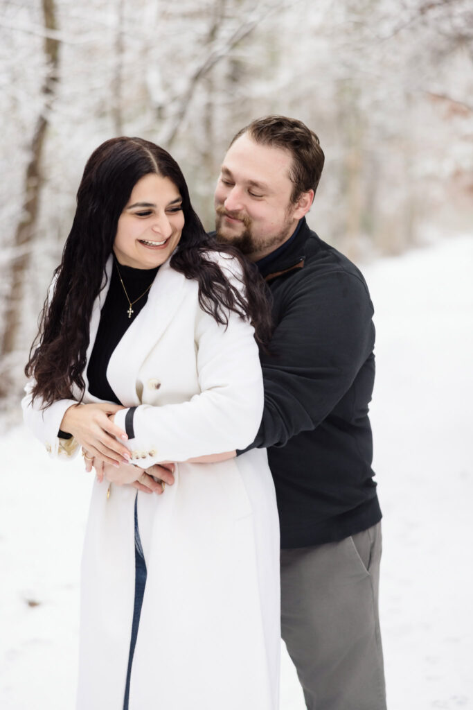 Engaged couple embracing in a snowy path, with the bride-to-be smiling in a white winter coat and the groom-to-be holding her from behind in a quiet, intimate moment.