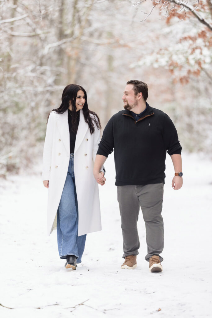Engaged couple walking hand-in-hand through a snow-covered path at Bethpage State Park, sharing a warm smile in a quiet winter landscape.