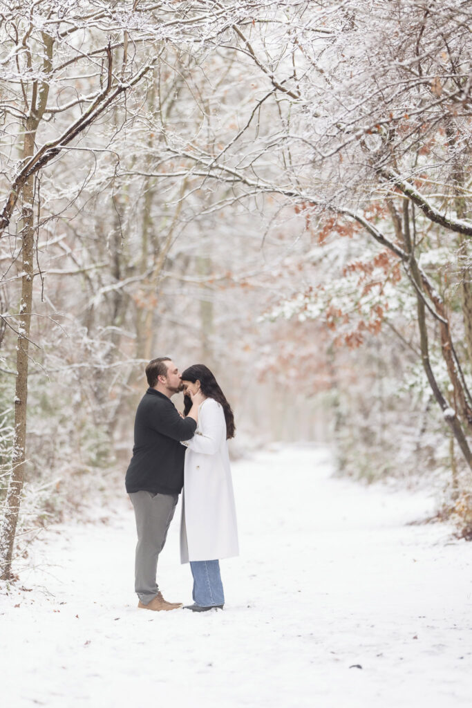 Groom-to-be kisses his fiancée’s forehead in a snow-covered woodland path at Bethpage State Park, framed by delicate winter branches and quiet intimacy.