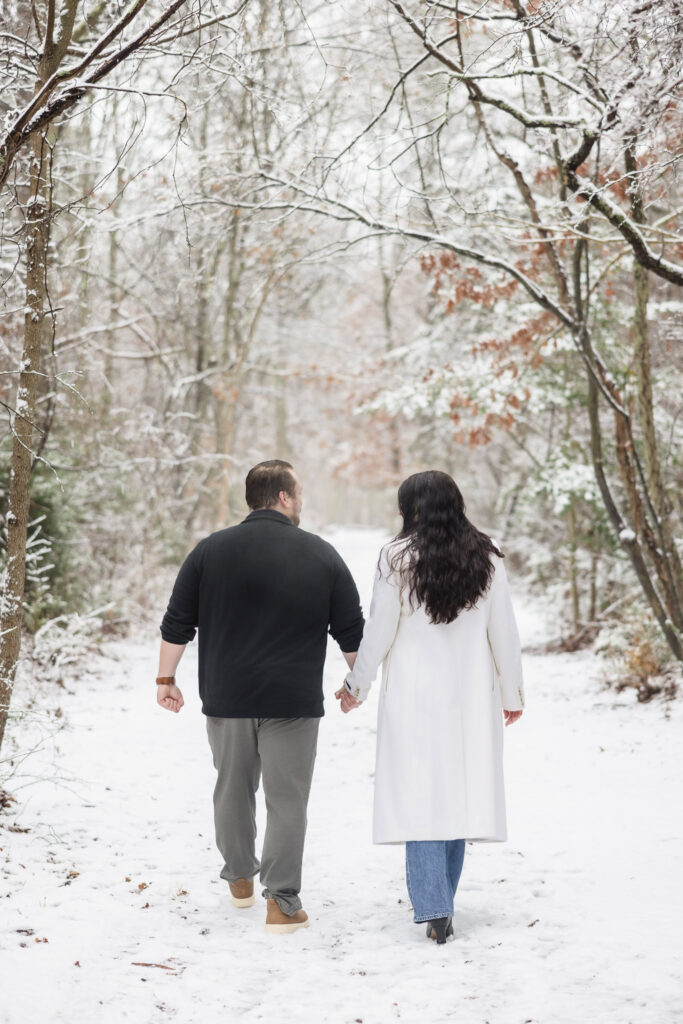 Couple walking hand-in-hand through a snowy forest trail at Bethpage State Park, captured from behind to highlight their quiet connection in winter surroundings.
