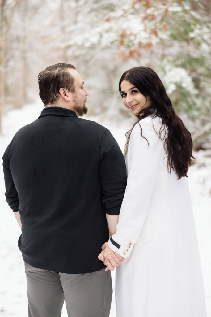 Bride-to-be looking over her shoulder with a soft smile while holding hands with her fiancé during their snowy engagement session.