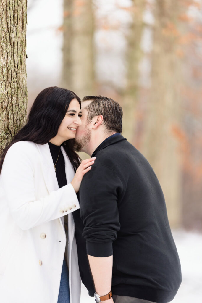 Groom-to-be whispers into his fiancée’s ear as she laughs, leaning against a tree during their snowy winter engagement session.