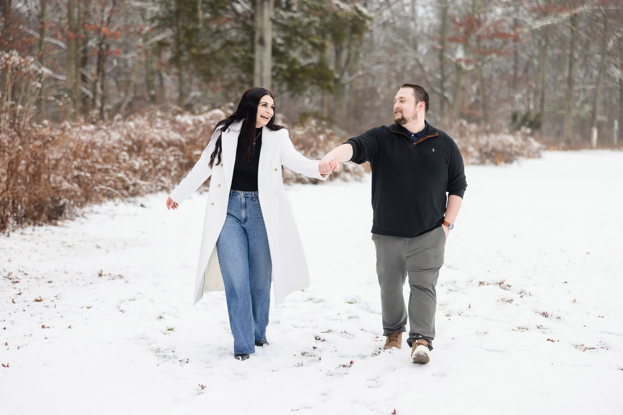 Couple holding hands and walking through a snowy open field at Bethpage State Park, smiling and enjoying a playful winter engagement session.