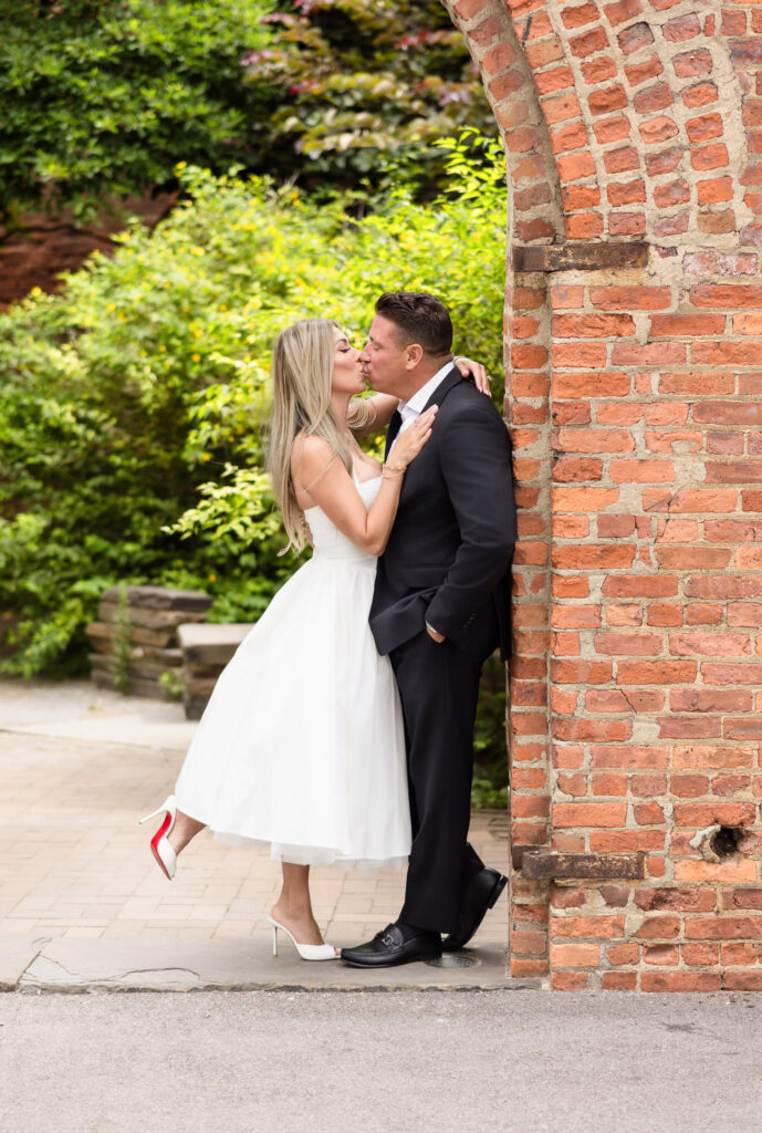 Couple kissing beneath a historic brick arch in DUMBO Brooklyn, highlighting one of the best photo locations in DUMBO for engagement photos with classic red brick textures and romantic garden surroundings.