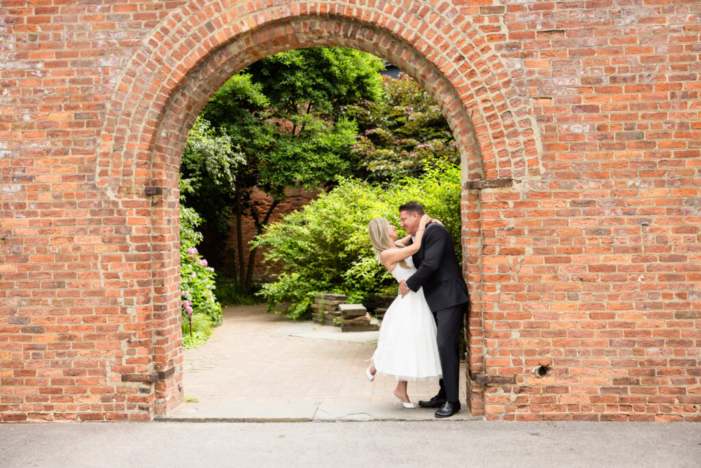 Couple in structured white dress and tailored black suit embracing beneath a brick archway in DUMBO Brooklyn, demonstrating what to wear for engagement photos in DUMBO against classic architectural backdrops and cobblestone textures.