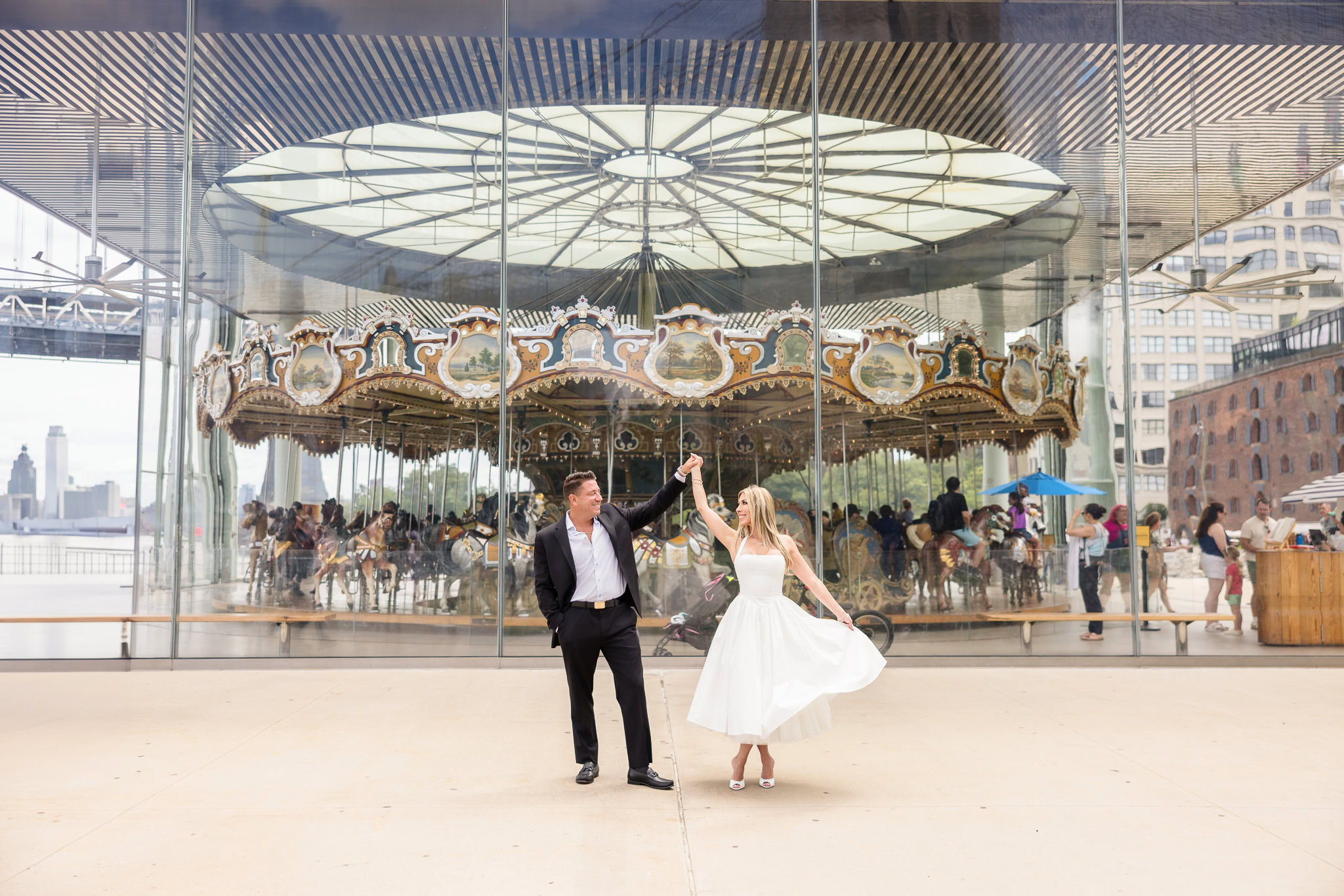Couple dancing in formal NYC engagement attire in front of Jane’s Carousel in DUMBO Brooklyn, showcasing structured silhouettes and refined styling against a glass architectural backdrop.