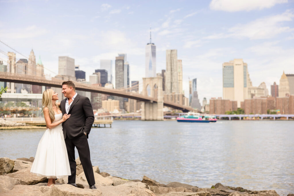 Couple in formal engagement attire NYC standing along the Brooklyn waterfront in DUMBO with the Manhattan Bridge and skyline behind them, illustrating what to wear for engagement photos in DUMBO with neutral tones and structured silhouettes.