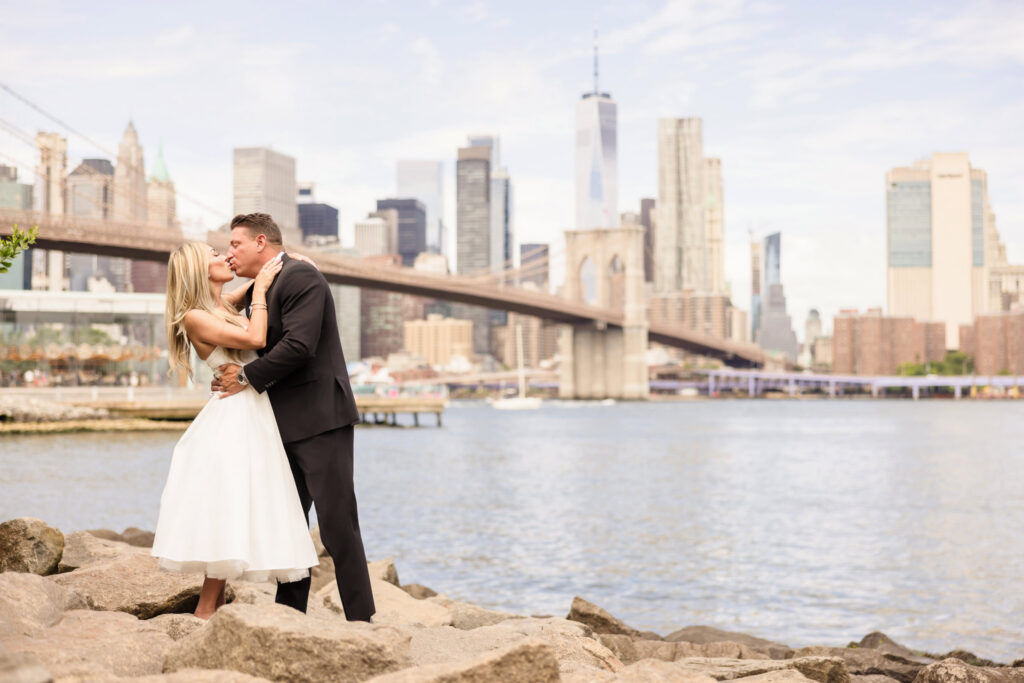 Couple embracing on the rocks at Pebble Beach Brooklyn with the Brooklyn Bridge and Manhattan skyline behind them, showcasing one of the best photo locations in DUMBO for engagement photos along the Brooklyn waterfront.