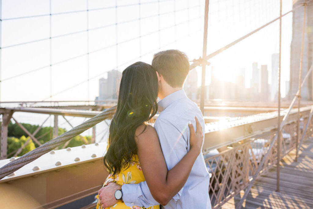 Brooklyn Bridge engagement photos in DUMBO capturing Sreetha and Demitri embracing at golden hour, with warm evening light in Brooklyn illuminating the bridge cables and Manhattan skyline beyond.