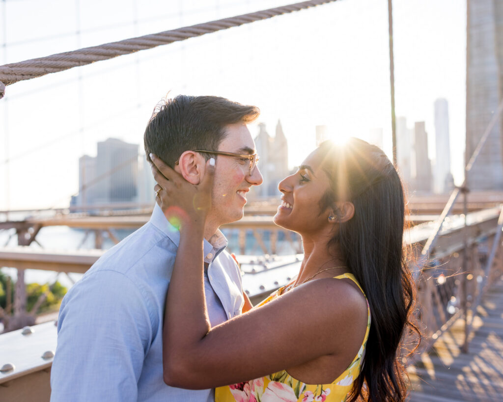 Close-up Brooklyn Bridge engagement photos in DUMBO of Sreetha and Demitri smiling at golden hour, with sun flare through the bridge cables and the Manhattan skyline creating warm evening light in Brooklyn.