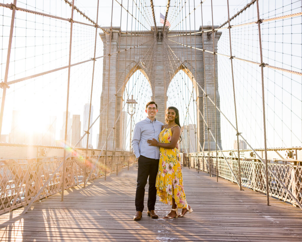 Full-length portrait of couple centered on the Brooklyn Bridge at golden hour, showcasing iconic architectural engagement photos in NYC with symmetrical cables and skyline views.