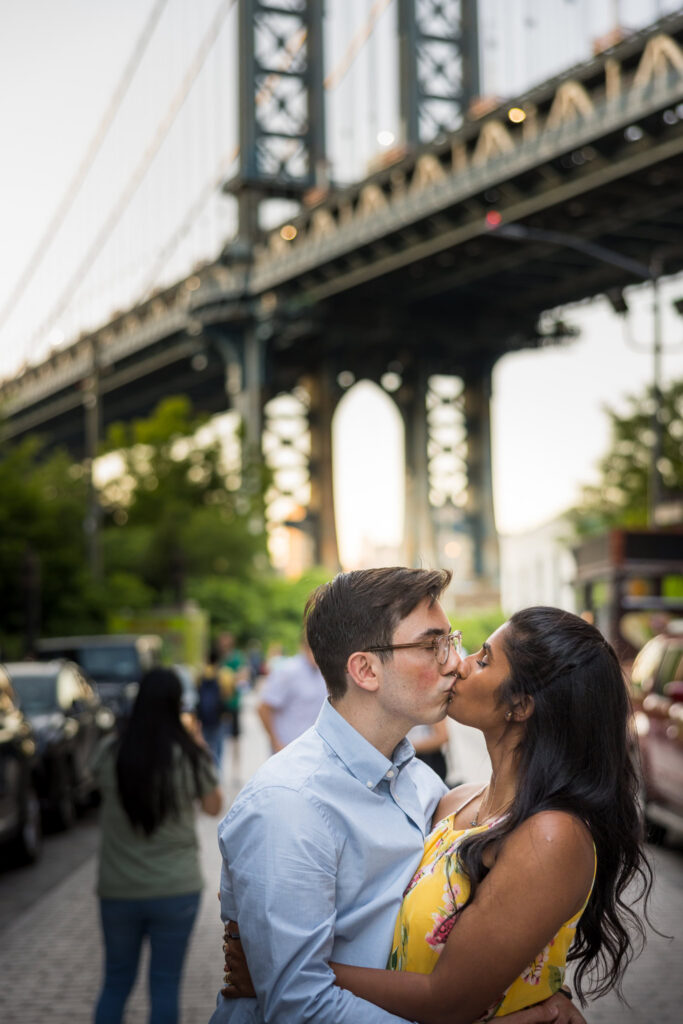 Manhattan Bridge engagement photos on Washington Street in DUMBO, featuring Sreetha and Demitri kissing beneath the bridge with cobblestone streets and architectural framing in Brooklyn.