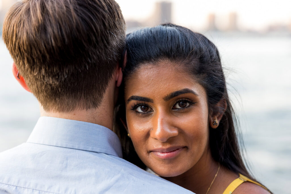 Close-up NYC couple portrait photos, featuring Sreetha resting against Demitri with soft waterfront light and a blurred Brooklyn skyline backdrop.