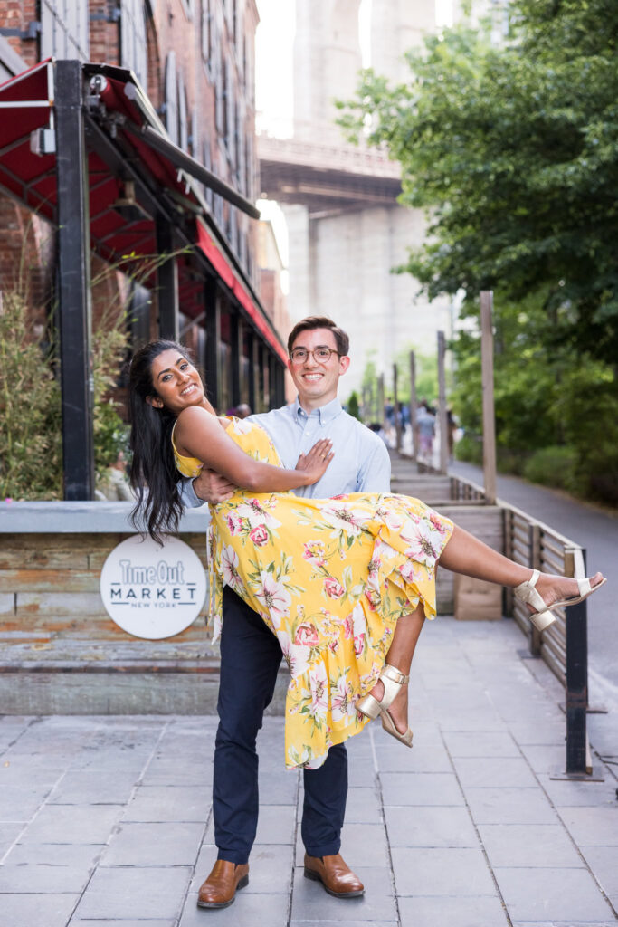 Playful DUMBO engagement session portrait near Time Out Market, with Demitri lifting Sreetha in a yellow floral dress beneath the Manhattan Bridge and brick-lined cobblestone streets in Brooklyn.