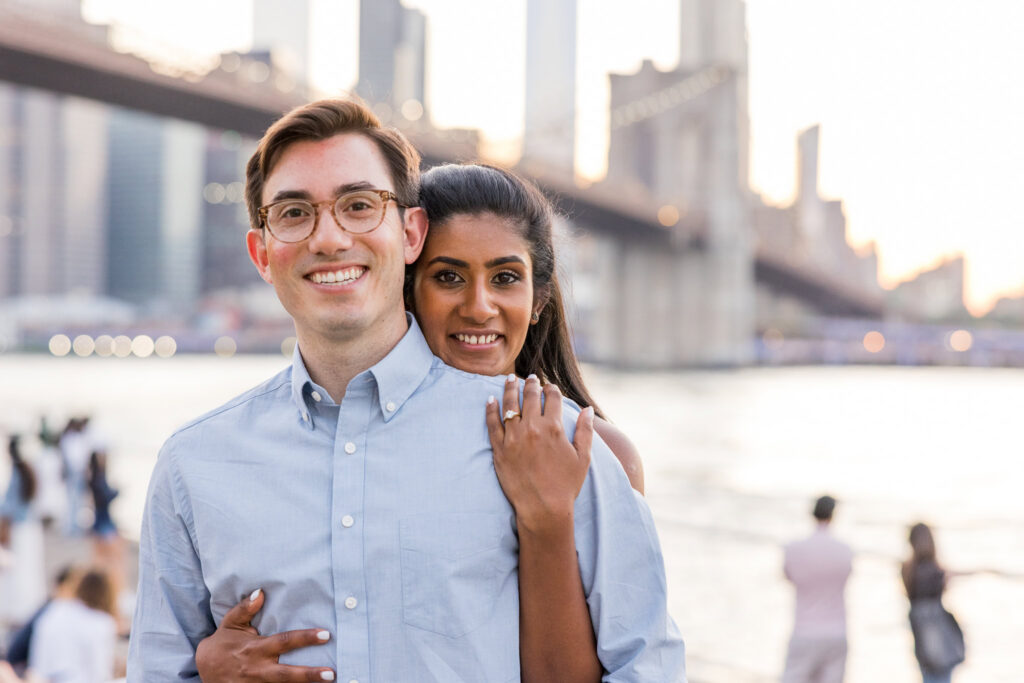 Brooklyn Bridge Park engagement photo at golden hour, with Sreetha and Demitri embracing along the waterfront and the Brooklyn Bridge and Lower Manhattan skyline softly glowing behind them.