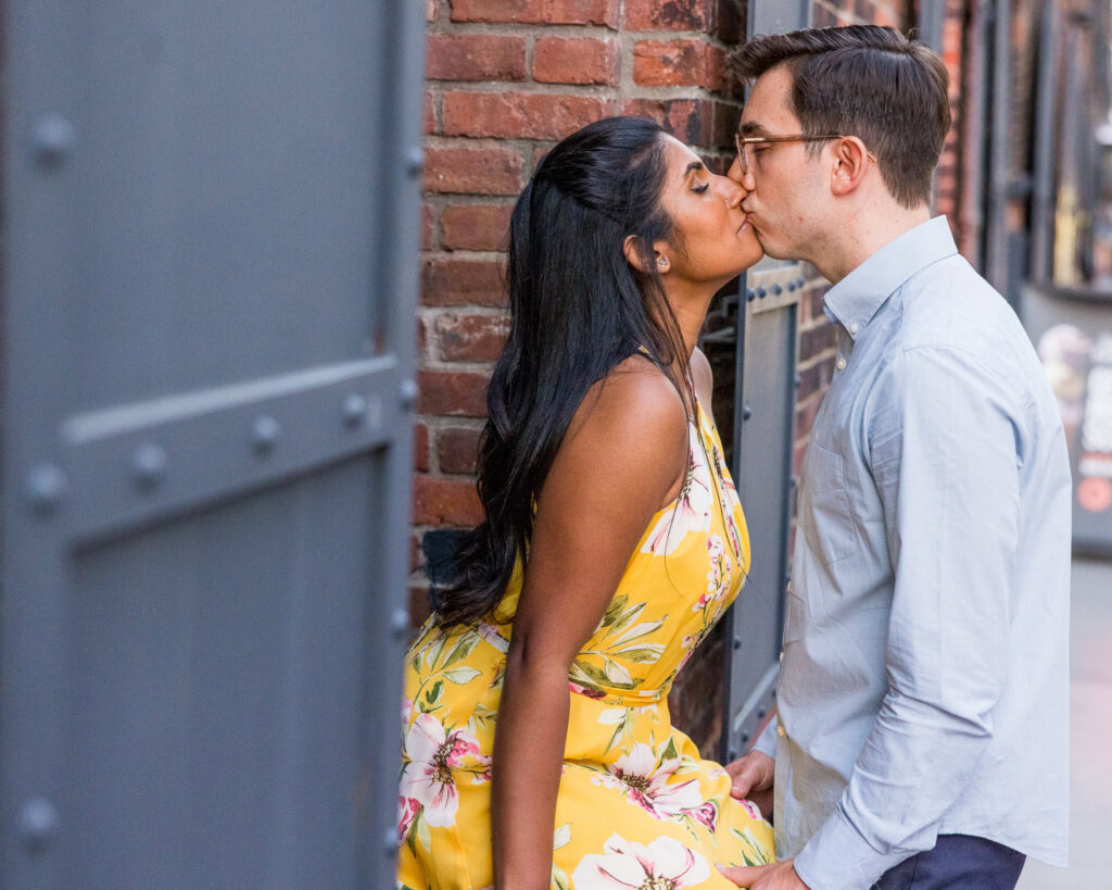 Intimate Brooklyn Bridge engagement photo in DUMBO, with Sreetha and Demitri kissing against a brick wall and industrial metal doors for a refined architectural NYC backdrop.
