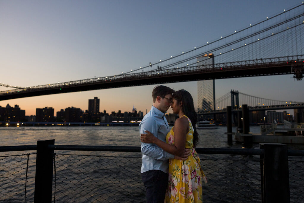 Evening Brooklyn Bridge Park engagement photo with Sreetha and Demitri embracing at blue hour, Manhattan Bridge illuminated above the East River and NYC skyline glowing in the background.